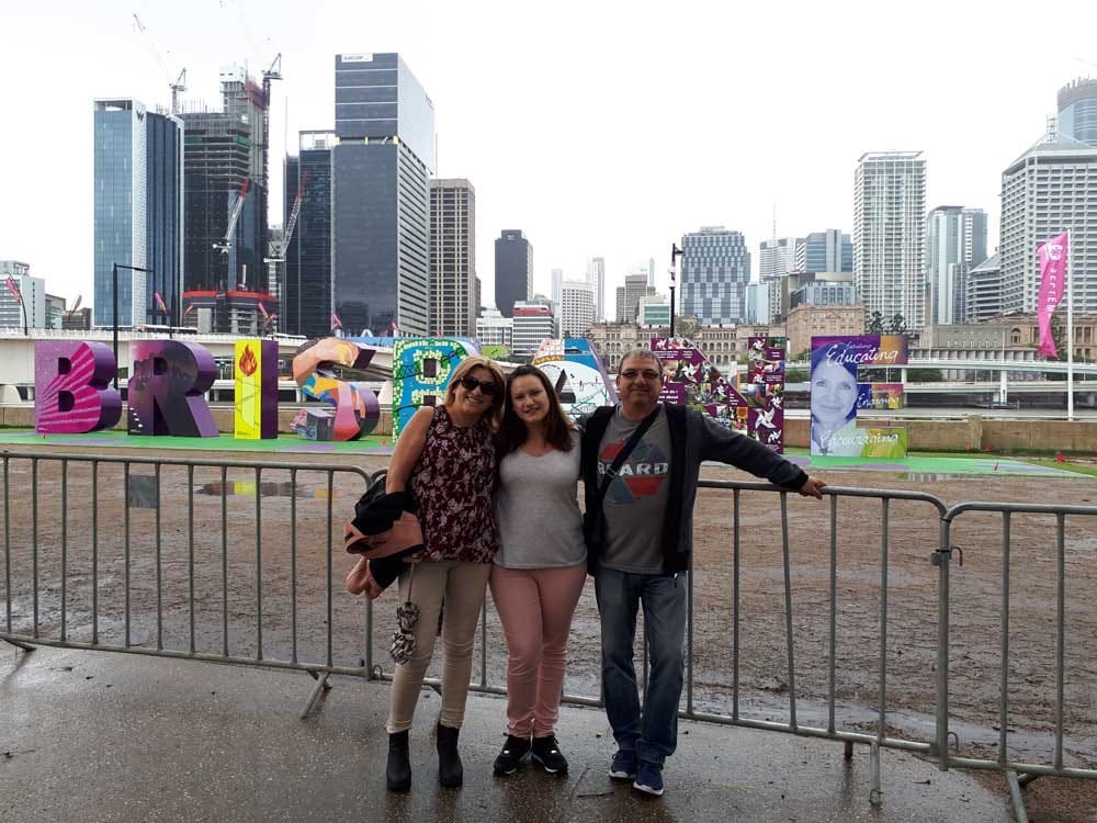 chica morena con los padres posando para cámara delante de las letras de la ciudad de Brisbane. Al fondo se ven los rascacielos de la ciudad